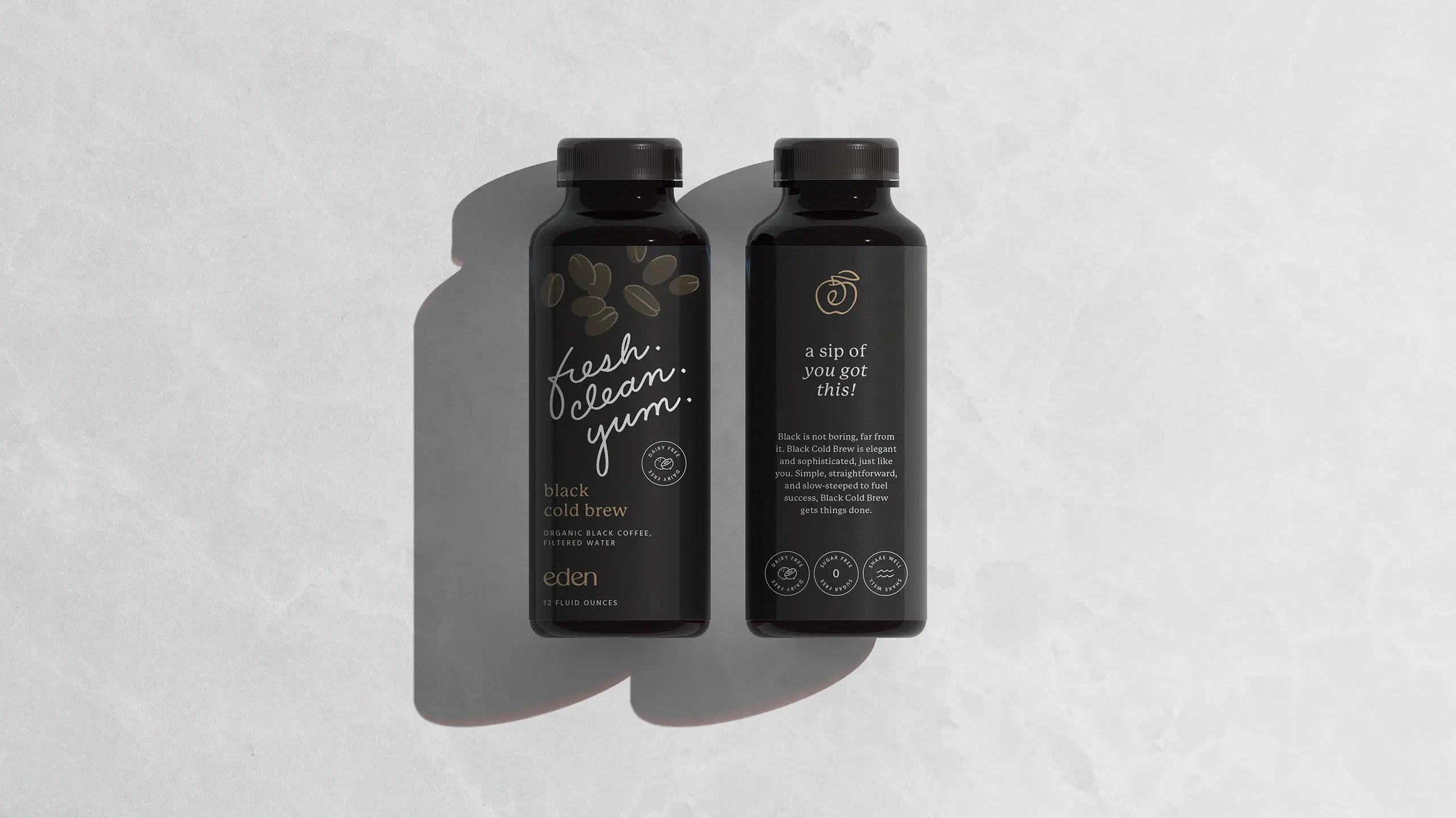 Front and back of a black Eden cold brew bottle with fresh. clean. yum. in white script, on a light marble surface.