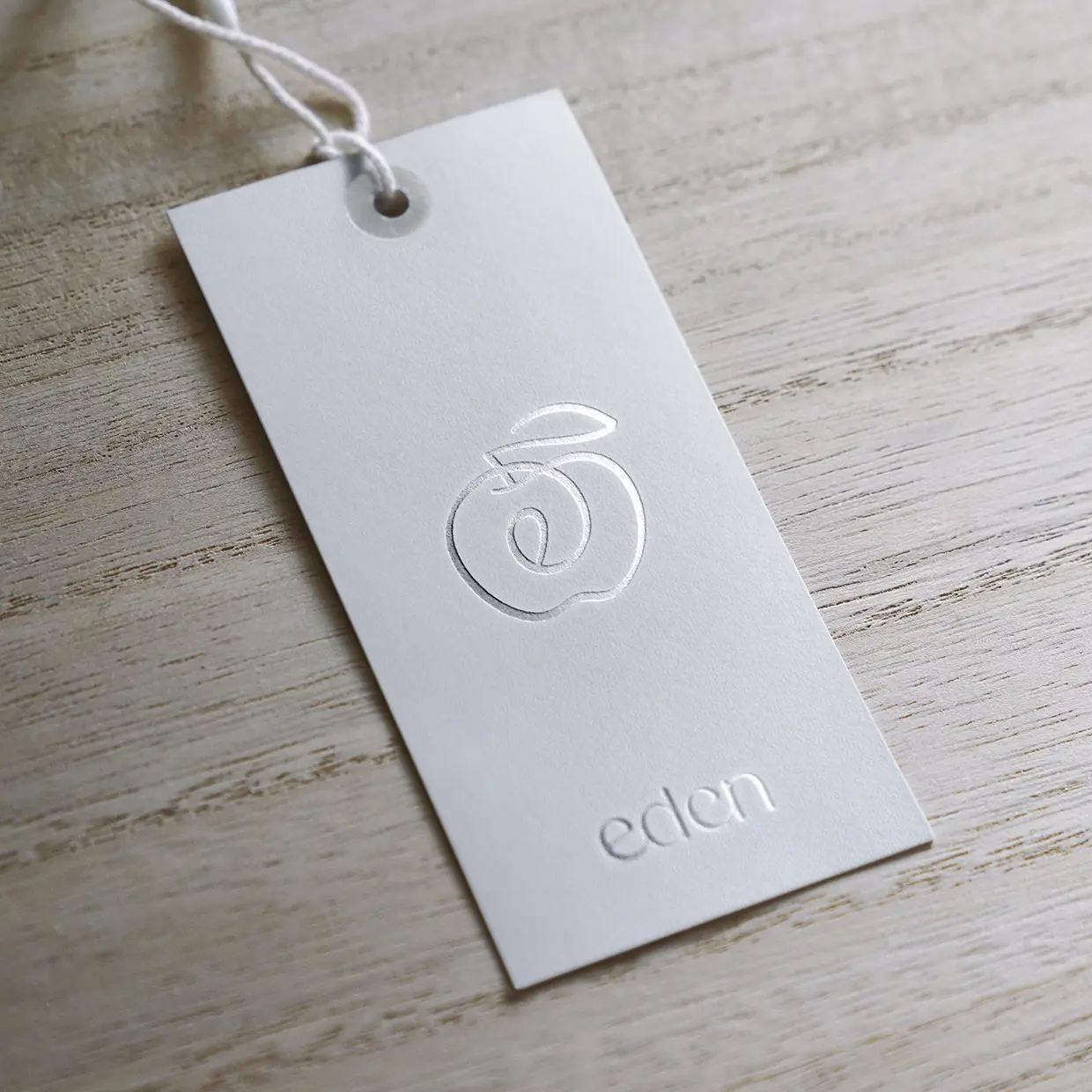 Eden hang tag with an embossed silver apple outline and the eden wordmark, tied with white string, on a light wood surface.