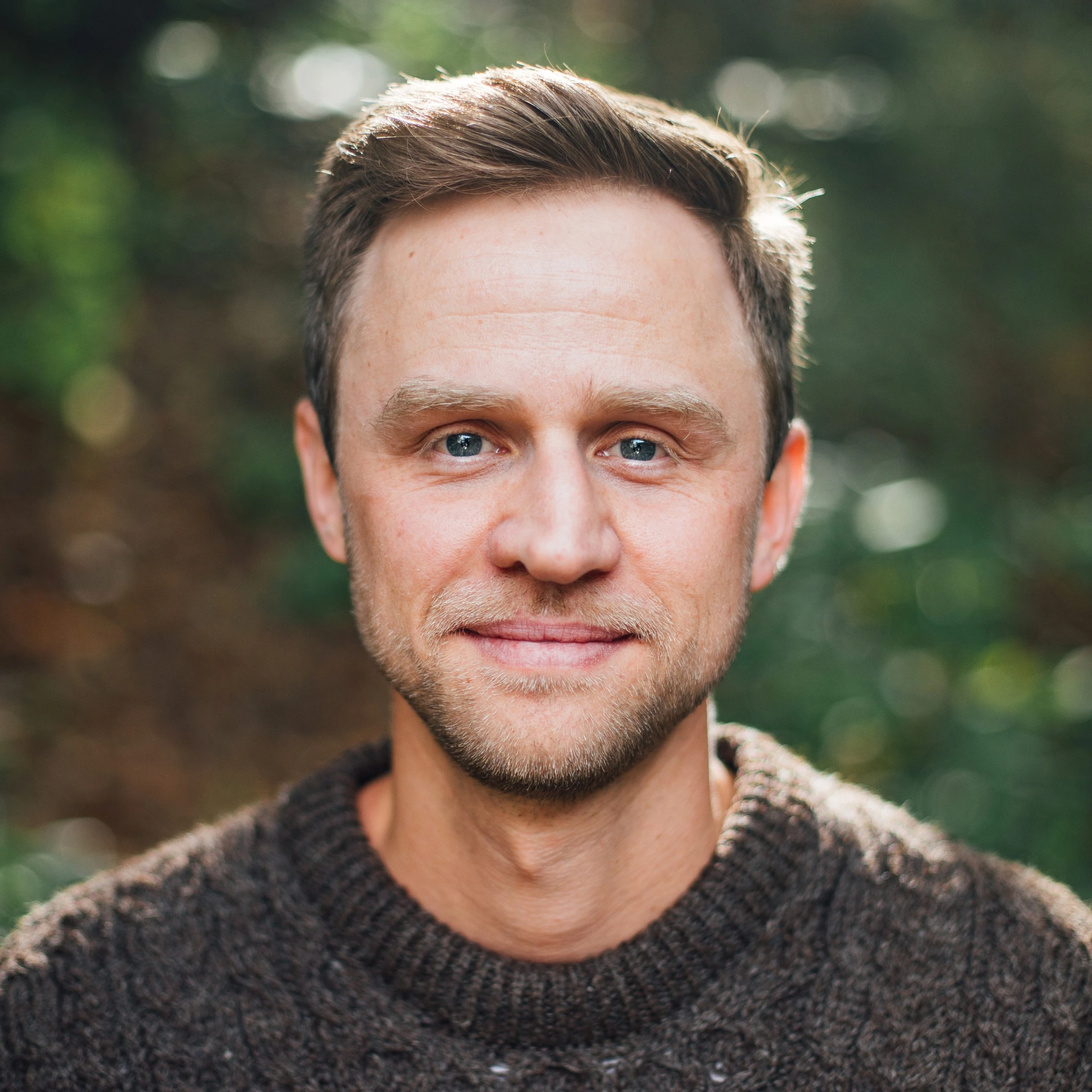Headshot of a man with short brown hair and light stubble in a brown knit sweater, against blurred green foliage.