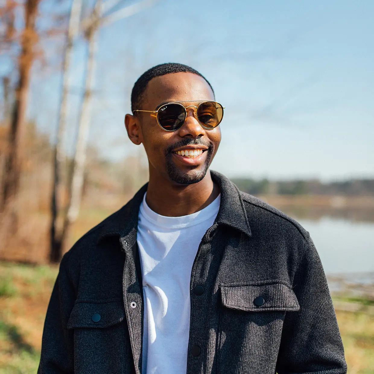 Smiling man in round gold sunglasses, a white T-shirt and a charcoal shirt jacket, standing by a lake on a sunny day.