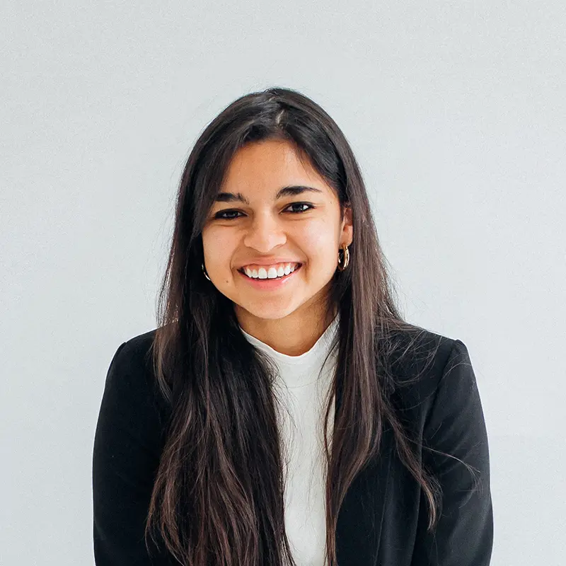 Portrait of Brenda Paquin smiling, with long dark hair, a black blazer and a white top, against a light gray wall.