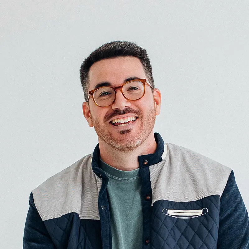 Portrait of John Howard smiling in tortoiseshell glasses and a navy and gray quilted jacket over a green T-shirt.