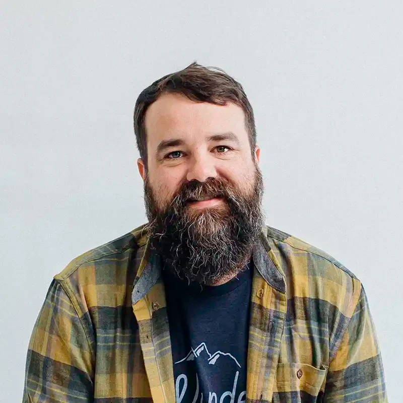Portrait of Josh Brown, with a full beard, in a green and navy plaid flannel shirt over a navy graphic T-shirt.