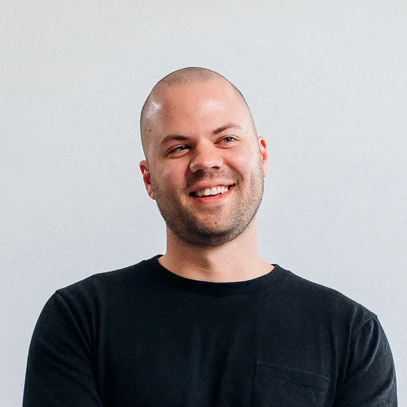 Portrait of Tyler Rees smiling in a black crew-neck T-shirt against a light gray wall.