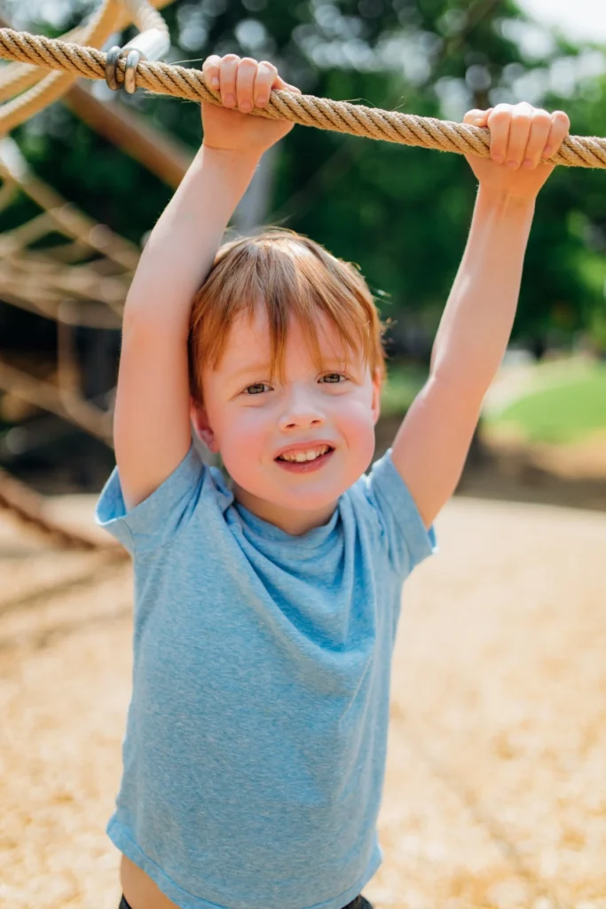 A red-haired boy in a blue T-shirt smiles as he hangs from a rope climber at a playground.