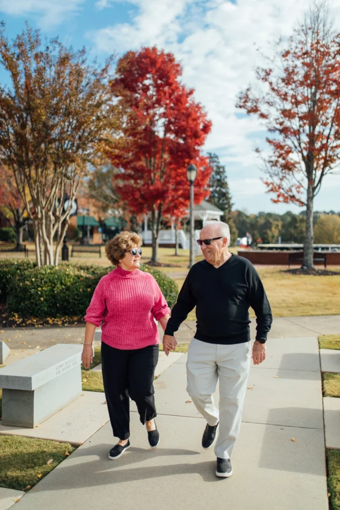 An older couple holds hands as they stroll through a downtown park under bright red autumn trees.