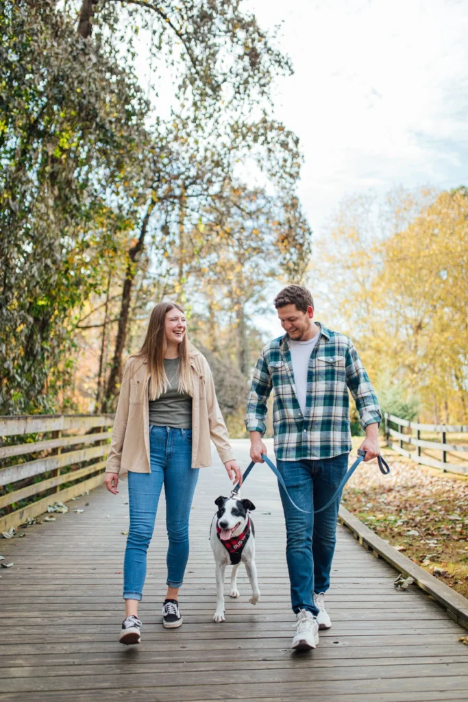 A young couple laughs as they walk a black and white dog along a wooden boardwalk lined with autumn trees.