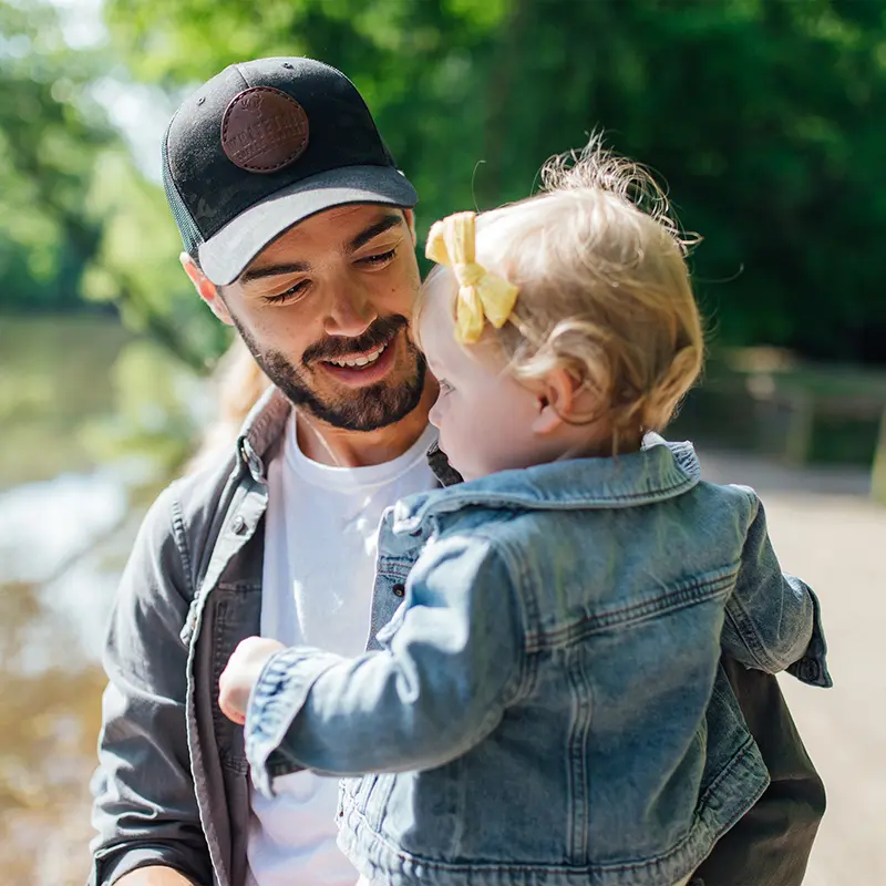 A smiling bearded man in a black cap holds a toddler in a denim jacket and yellow hair bow beside a wooded lake.