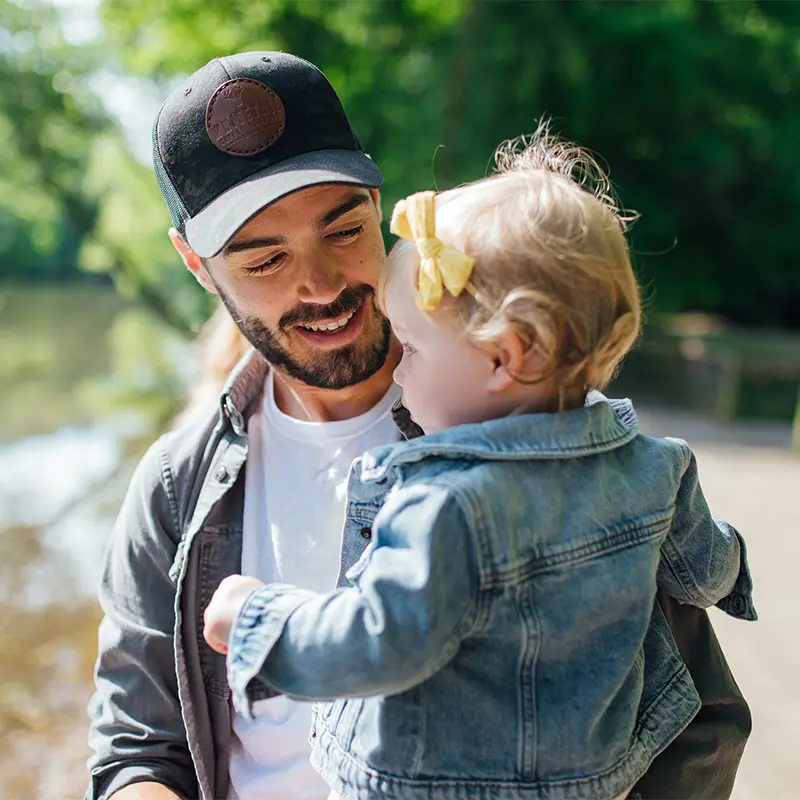 A smiling bearded man in a black cap holds a toddler in a denim jacket and yellow hair bow beside a wooded lake.