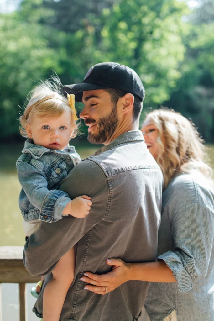 A father in a black cap holds his toddler daughter while the mother leans in smiling, beside a leafy lake.