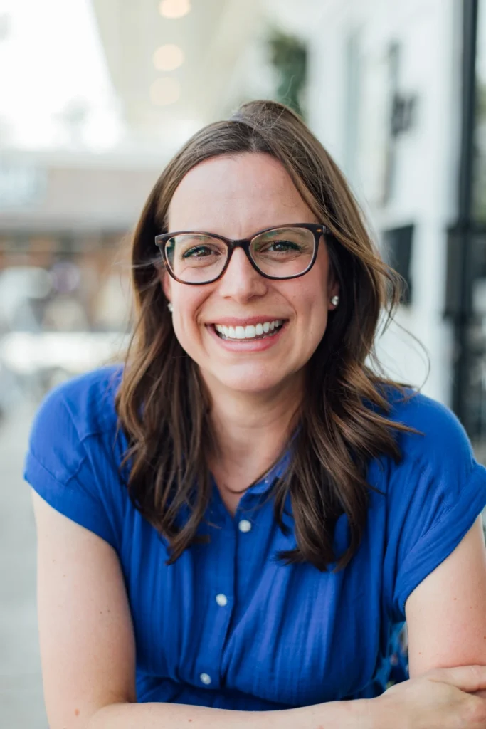 Smiling woman with dark-rimmed glasses and a blue blouse leans on a table at a sidewalk cafe.