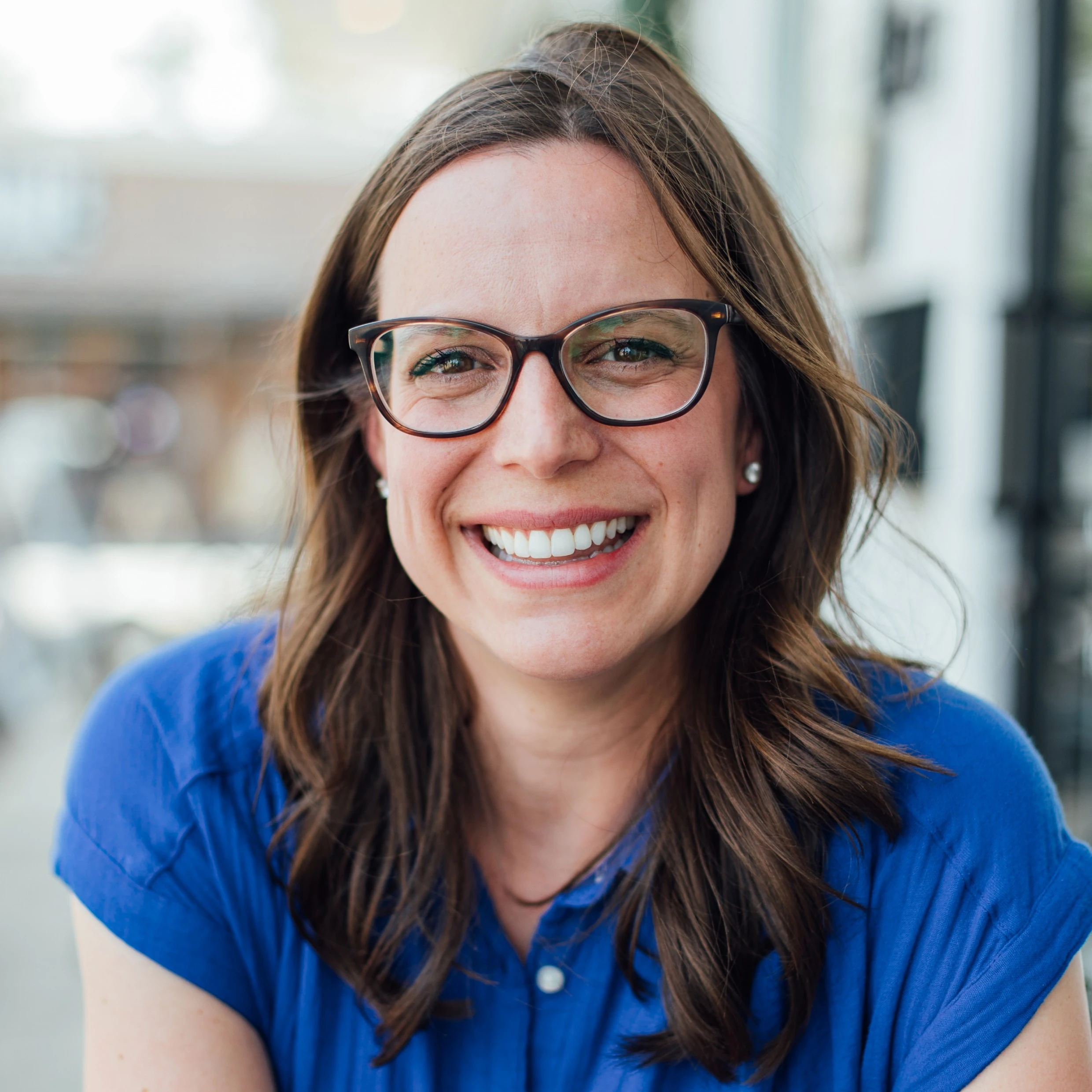 Smiling woman with shoulder-length brown hair and dark-rimmed glasses in a blue blouse on a sunny downtown sidewalk.